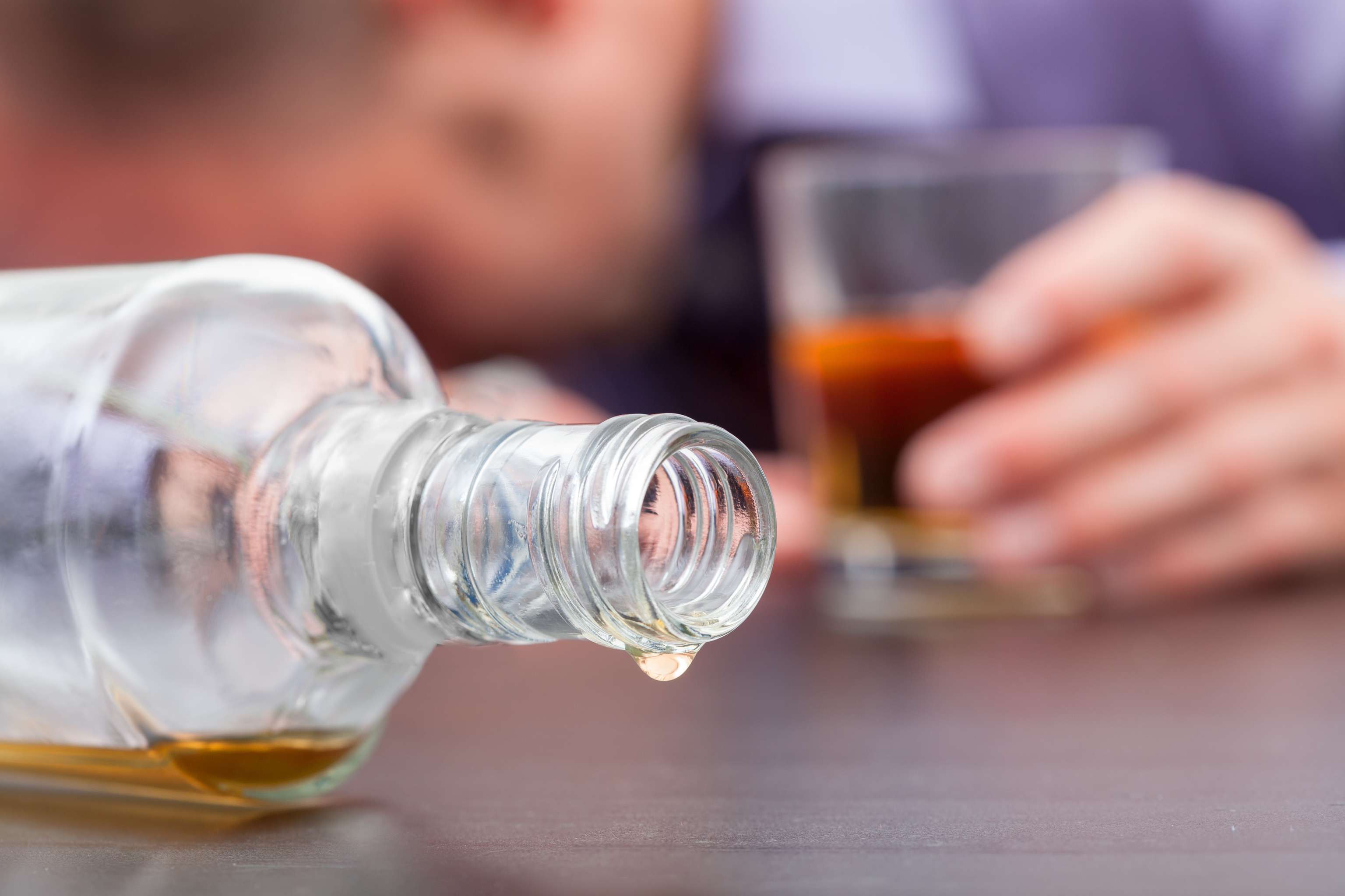 Consommation non contrôlée d'alcool Gros plan sur une bouteille de whisky renversée et presque vide avec, en arrière-plan, une personne endormie avec un verre de whisky à moitié plein.