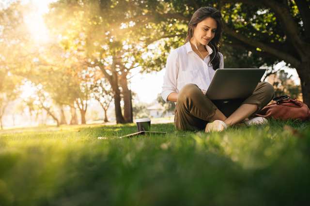 A woman works outside on a laptop with trees in the background. Woman sitting on grass in park working on laptop. Women wearing headphones with laptop while sitting under tree in park with bright sunlight from behind.