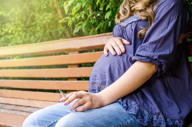 close up of pregnant woman smoking cigarette outdoor in park on nature background habit forbidden A pregnant woman sits on a park bench smoking a cigarette.