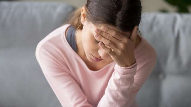 Young woman grabs her forehead. Young tired unhealthy mixed race woman sitting on sofa in living room at home with eyes closed, holding head with hand, suffering from severe sudden headache or migraine, whirling pain.