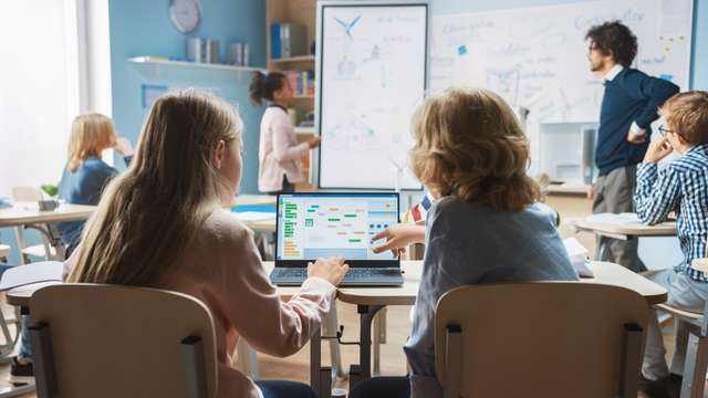 A class with two children in the foreground. They work with a laptop. Elementary School Science Class: Over the Shoulder Little Boy and Girl use laptop with Screen Showing Programming Software. Physics teacher explains lesson to a diverse class full of smart kids