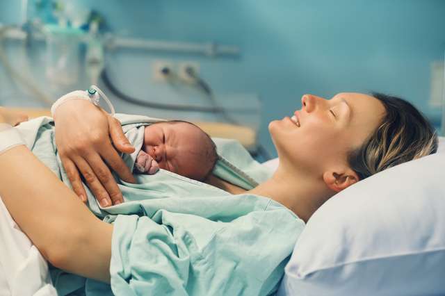 Mother and newborn. Birth of child in maternity hospital. Young mother hugging her newborn after birth. Women giving birth. First moments of baby life after labor. A mother is lying and holding her newborn child on her chest in a hospital, smiling with her eyes closed.