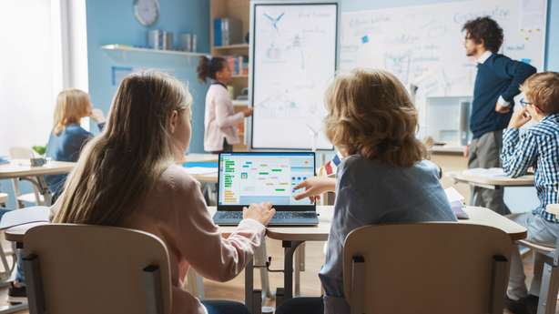 Une classe avec deux enfants au premier plan. Ils fonctionnent avec un ordinateur portable. Classe de sciences à l'école primaire : Un petit garçon et une petite fille en bandoulière utilisent un ordinateur portable avec un écran montrant un logiciel de programmation. Un professeur de physique explique une leçon à une classe diversifiée d'enfants intelligents.