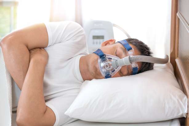 Hombre mayor feliz y saludable con máscara Cpap durmiendo toda la noche sobre su lado izquierdo cruzando los brazos sin roncar.Terapia de apnea obstructiva del sueño. Un señor mayor lleva una máscara respiratoria mientras está tumbado de lado en la cama intentando conciliar el sueño.