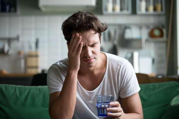 Jeune homme souffrant de maux de tête, de migraines ou de gueule de bois à la maison Photo d'un jeune homme avec des douleurs à la tête et un verre d'eau à la main
