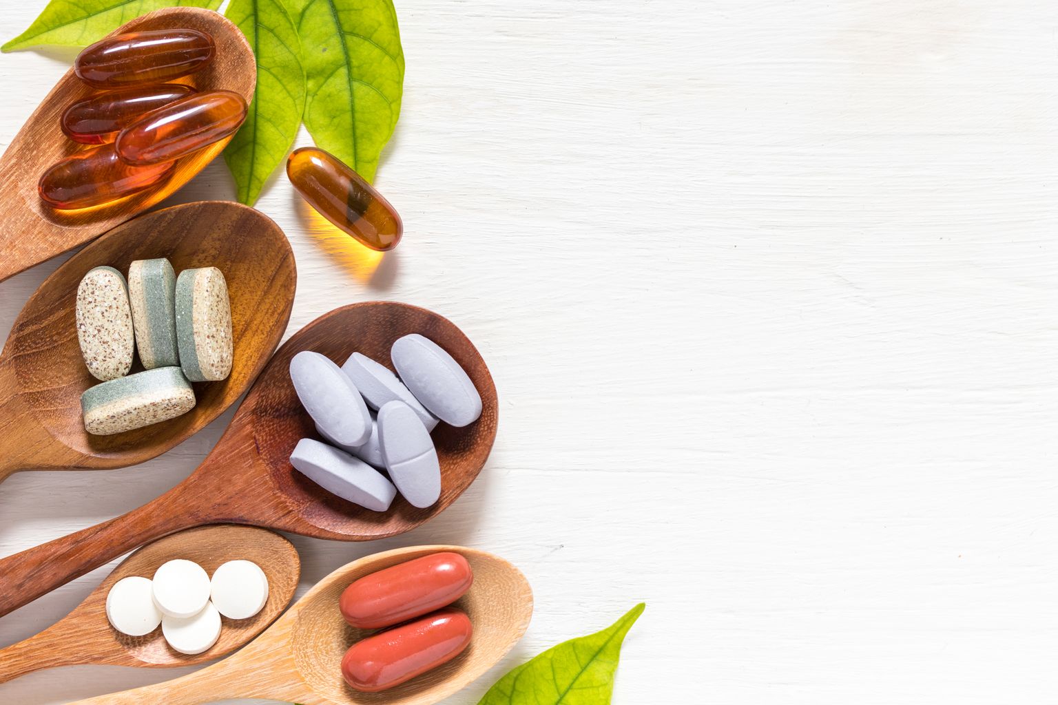 Variedad de píldoras de vitaminas en cucharas de madera sobre fondo blanco con hoja verde, producto complementario y médico, superficie plana de lego