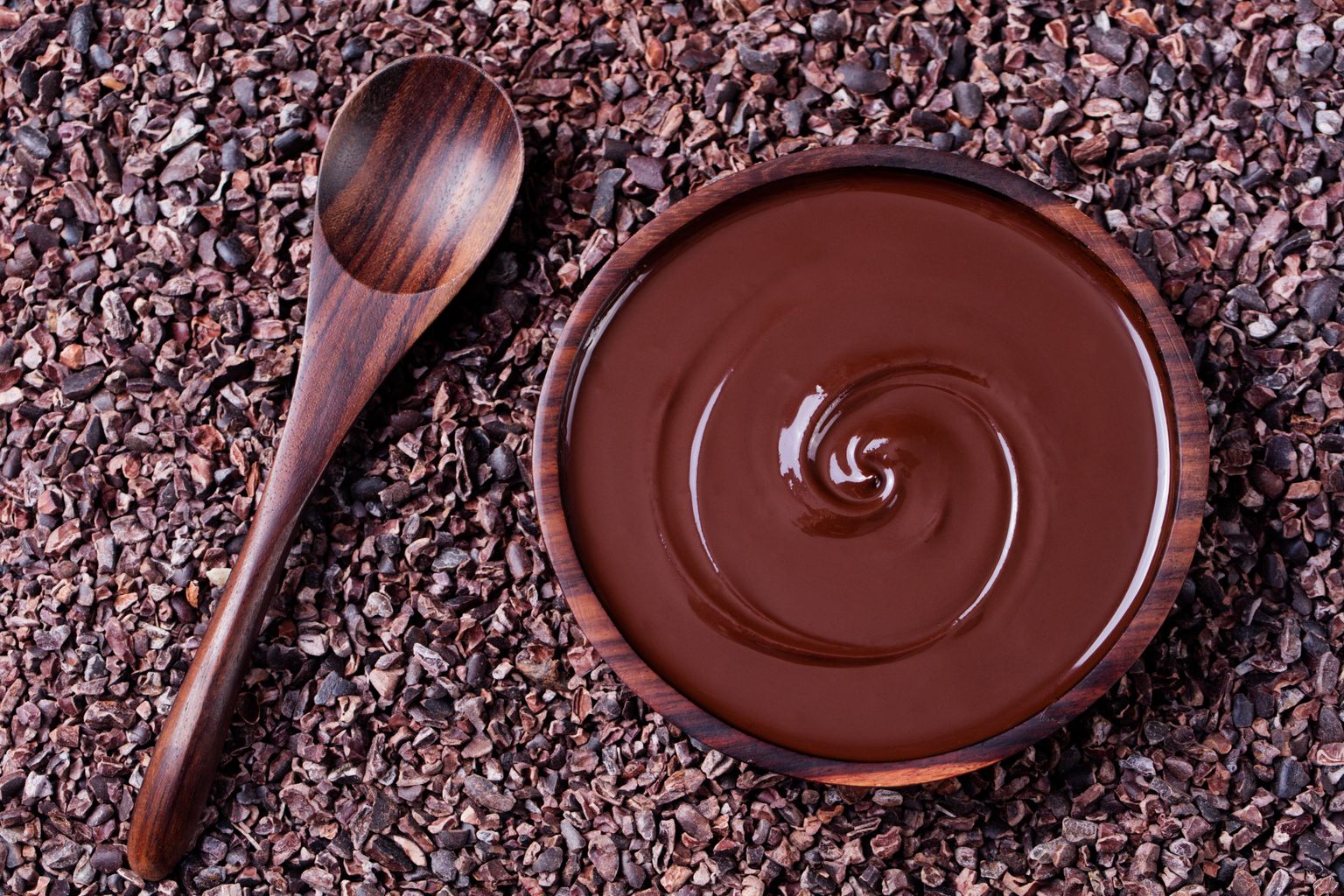 Ciotola di cioccolato fuso e cucchiaio di legno su uno sfondo di fave di cacao crude schiacciate, pennini. Spazio per la copia Vista dall'alto